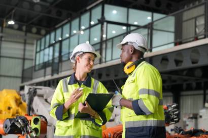 Image of two industrial workers wearing safety helmets and high-visibility jackets discussing a task with a clipboard inside a modern manufacturing facility.