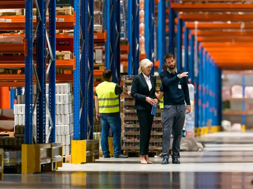 Image of several workers in a warehouse, wearing safety vests and helmets, standing among tall shelves filled with stacked boxes and pallets.