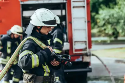 Image of a male firefighter in uniform and helmet, shown in close focus with a serious expression while standing near blurred emergency lights.