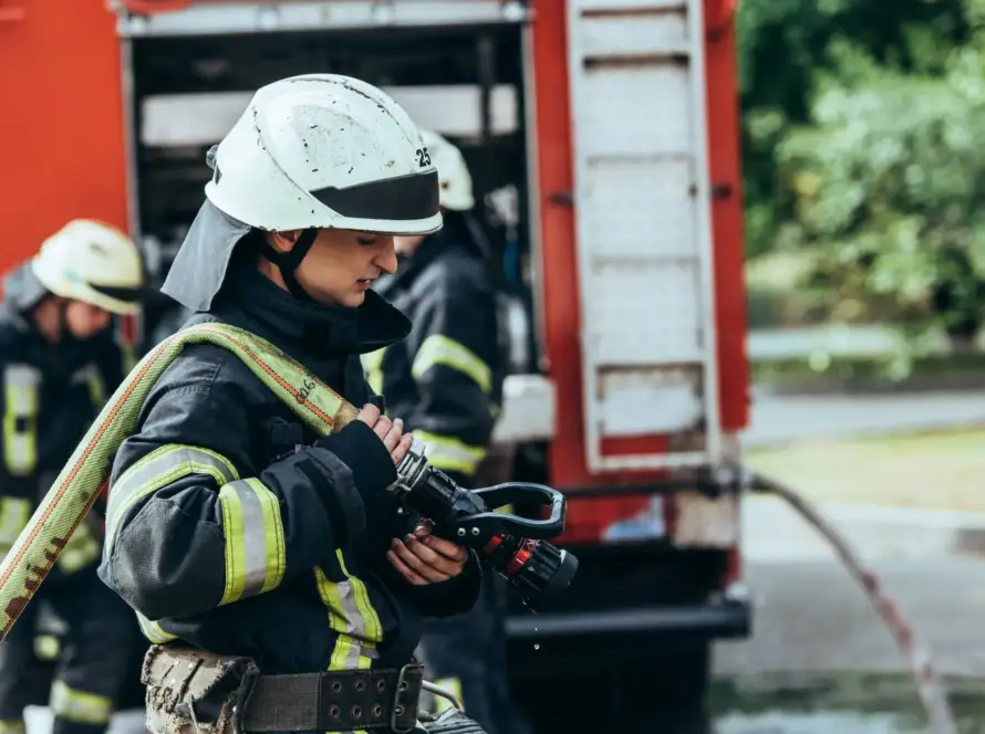 Image of a male firefighter in uniform and helmet, shown in close focus with a serious expression while standing near blurred emergency lights.