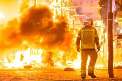 Image of a firefighter silhouette standing against bright flames, aiming a hose to spray water while battling a large, intense fire at night.