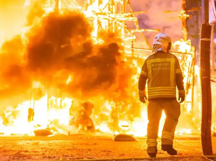 Image of a firefighter silhouette standing against bright flames, aiming a hose to spray water while battling a large, intense fire at night.