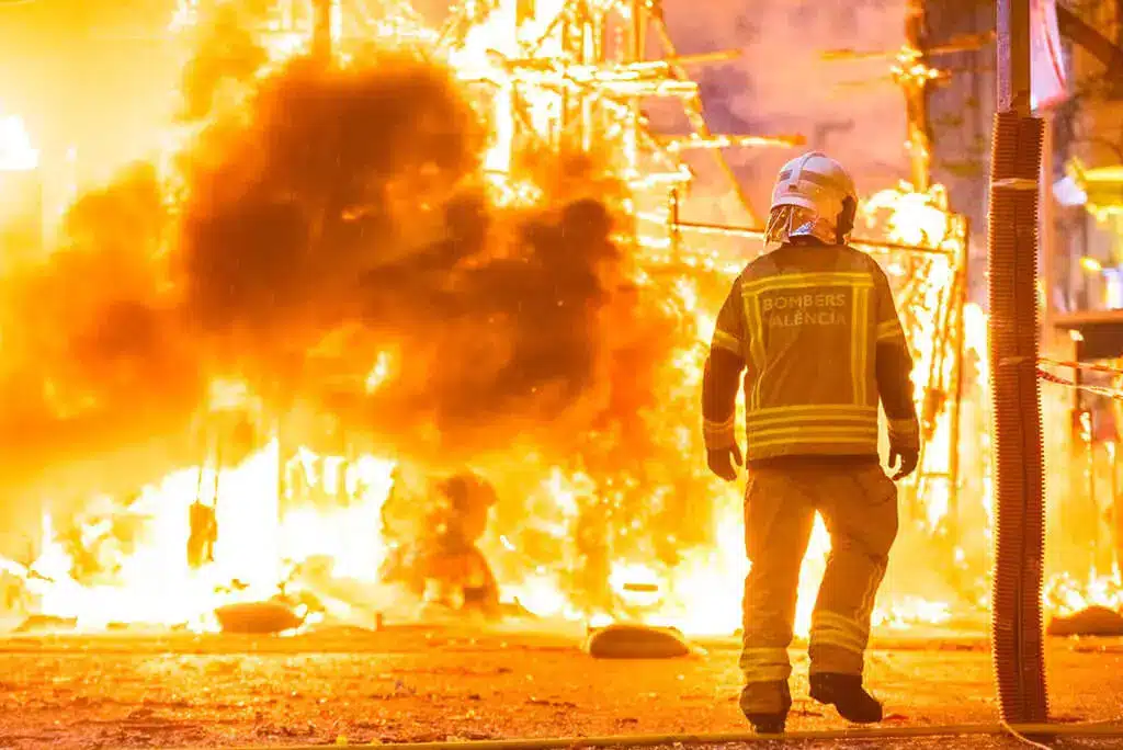 Image of a firefighter silhouette standing against bright flames, aiming a hose to spray water while battling a large, intense fire at night.