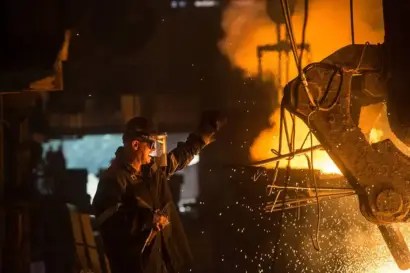 Image of a steelworker in protective gear standing near large industrial tanks, monitoring glowing hot metal during an active smelting process.