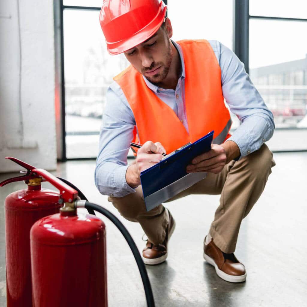Image of a fire risk assessment auditor in protective helmet and orange vest writing notes on a clipboard next to two fire extinguishers in a modern building