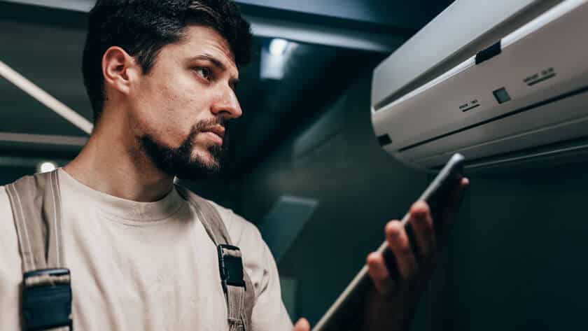 Image of a technician inspecting a wall-mounted air conditioning unit while holding a digital tablet in a modern indoor setting.