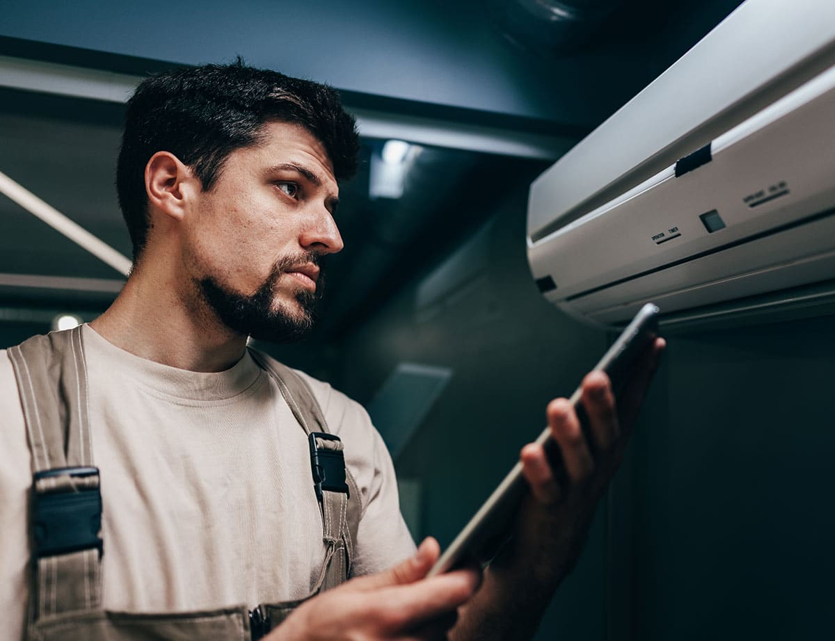 Image of a technician inspecting a wall-mounted air conditioning unit while holding a digital tablet in a modern indoor setting.