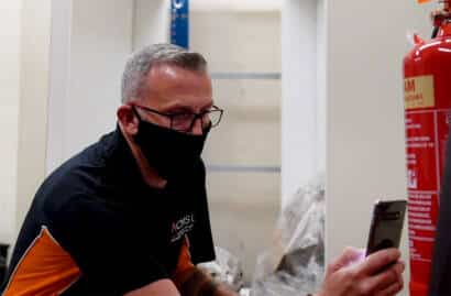 Image of a masked man inspecting a fire extinguisher as part of a Fire risk assessment checklist while holding a smartphone in a workshop setting.