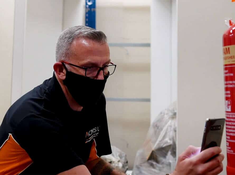 Image of a masked man inspecting a fire extinguisher as part of a Fire risk assessment checklist while holding a smartphone in a workshop setting.