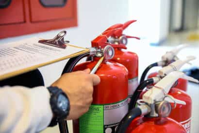 Image of a person inspecting a row of fire extinguishers while checking details against a clipboard during a safety audit.