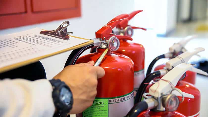 Image of a person inspecting a row of fire extinguishers while checking details against a clipboard during a safety audit.