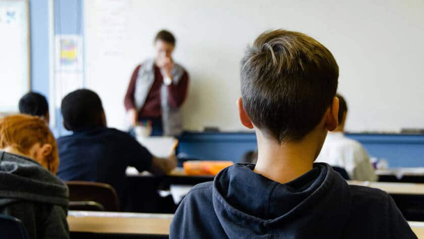 Image of a school classroom with a boy sat looking at the teacher in the distance.