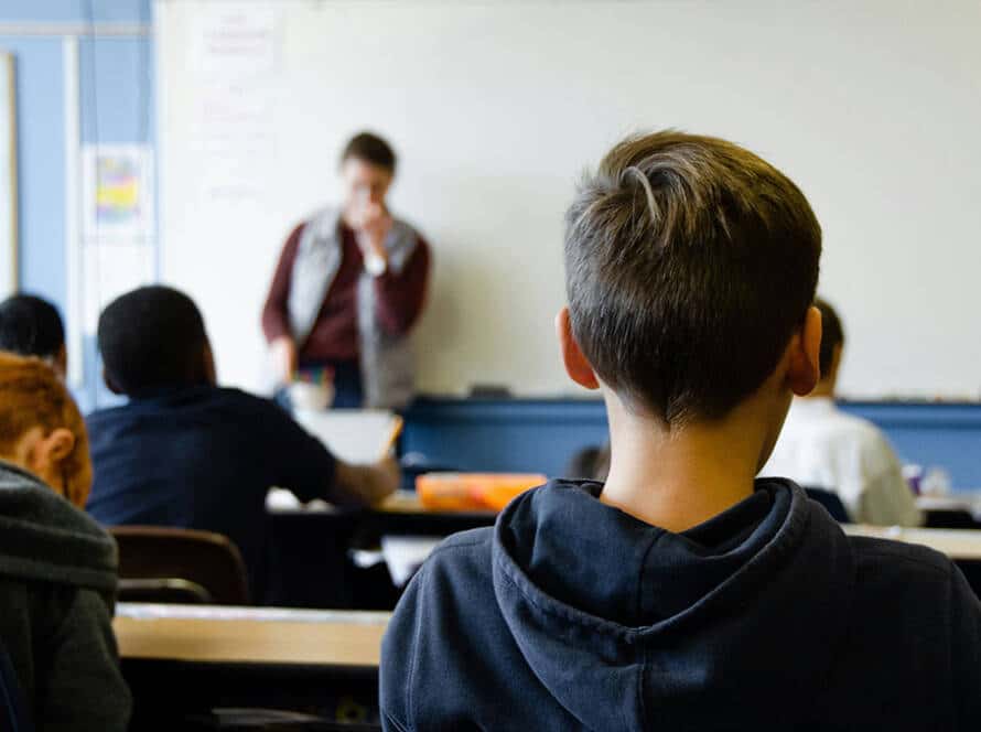 Image of a school classroom with a boy sat looking at the teacher in the distance.