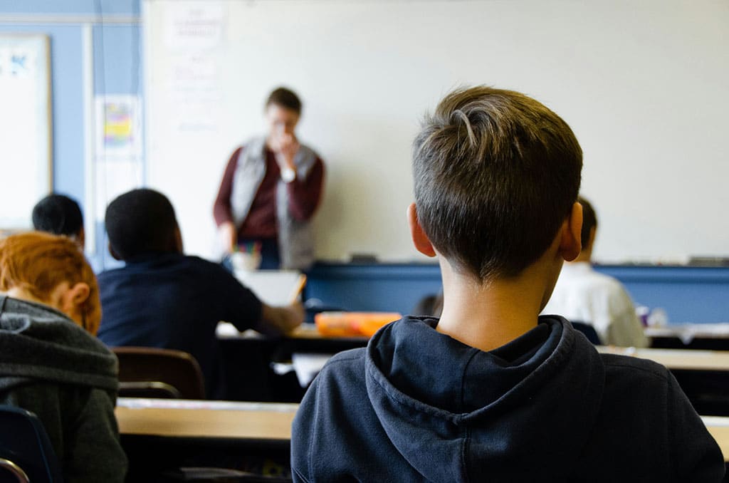 Image of a school classroom with a boy sat looking at the teacher in the distance.